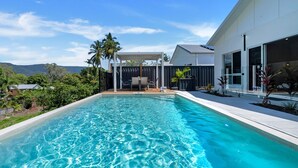Ein moderner Pool mit klarem, blauem Wasser, umgeben von einem weißen Gebäude mit großen Fenstern und einer überdachten Terrasse mit Gartenmöbeln.