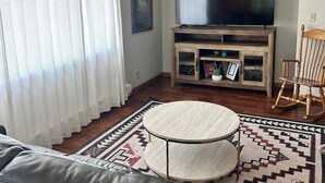 Ein Wohnzimmer mit einem Ledersofa, einem runden Couchtisch, einem Fernseher auf einem Holzständer, einem gemusterten Teppich und einem Holzschaukelstuhl.