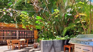 An outdoor patio with wooden tables and chairs, a barbecue grill, and lush greenery.