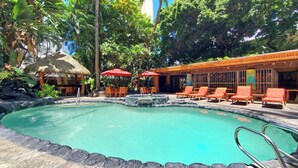 A pool area with lounge chairs and umbrellas, surrounded by tropical vegetation and a thatched-roof structure.