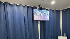 A room with dark blue curtains, a flat-screen TV mounted on the wall, a white pedestal, and a blue rug on the floor. - Golden Pond Cabins (Altmar)