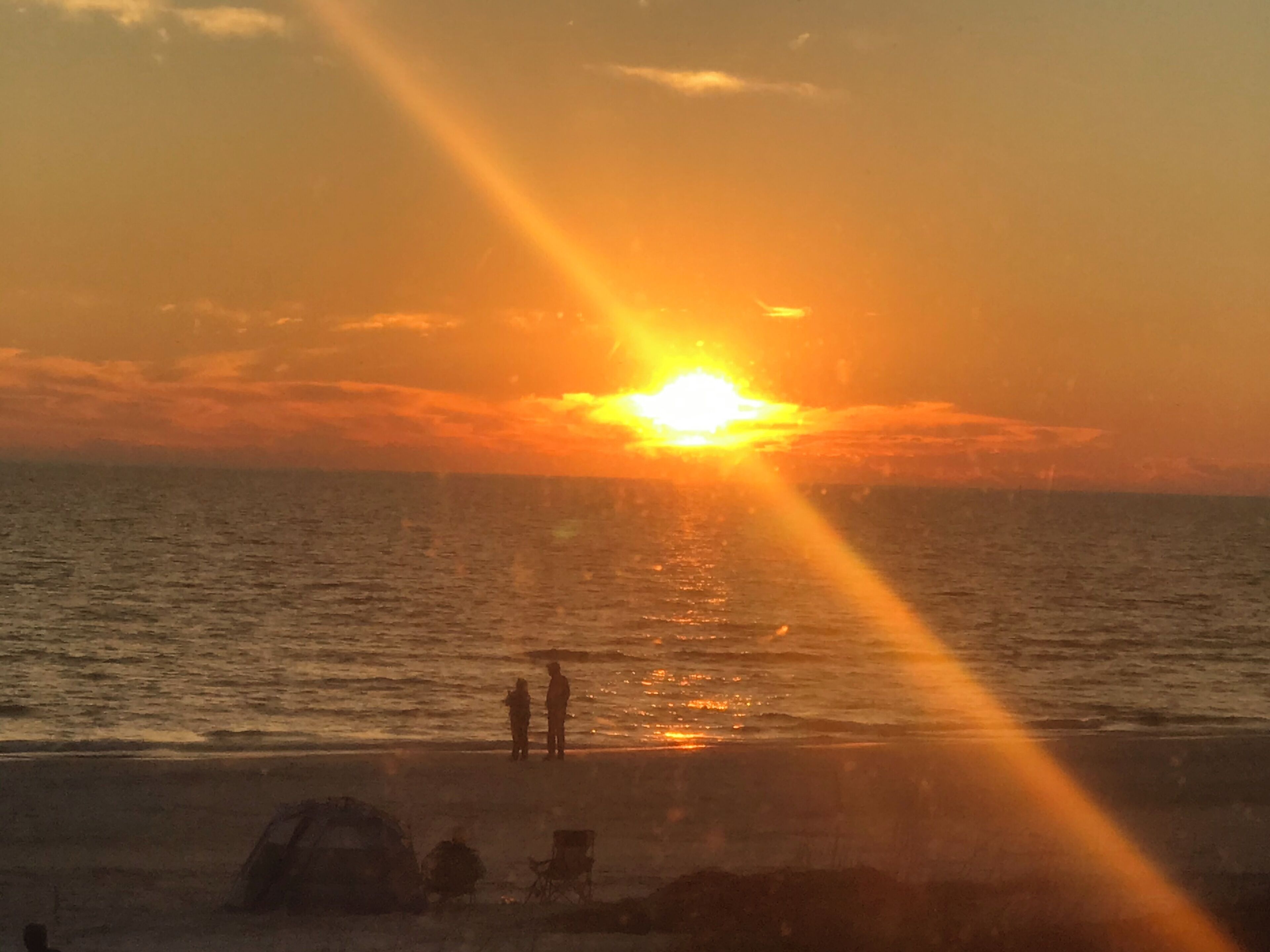Een zonsondergang boven de oceaan, met silhouetten van mensen en een tent op het strand.