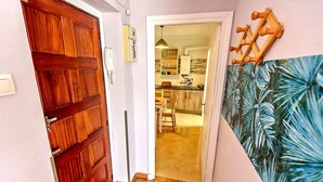 A hallway with a wooden door, a kitchen area visible through an open door, and a wall with a tropical leaf pattern.