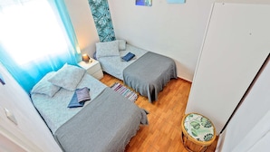 A small, cozy room with two beds, a wooden floor, and a window with sheer curtains.