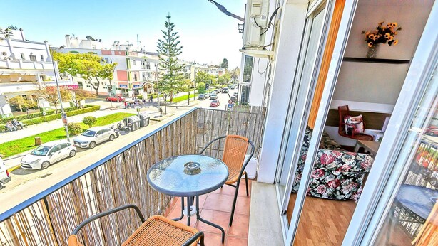 A balcony with wicker chairs, a small round table, and a view of a street with cars and buildings.
