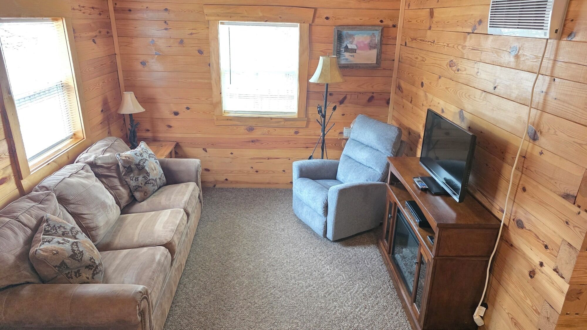 Habitación tipo cabaña de madera con sofá, sillón, televisor, lámpara y ventanas.