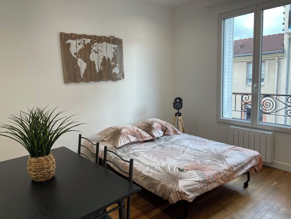 A bedroom with a bed, a small table with a plant, a world map on the wall, and a window with a view of buildings. - Newly renovated apartment in brilliant white—an oasis of tranquility. (Pantin)