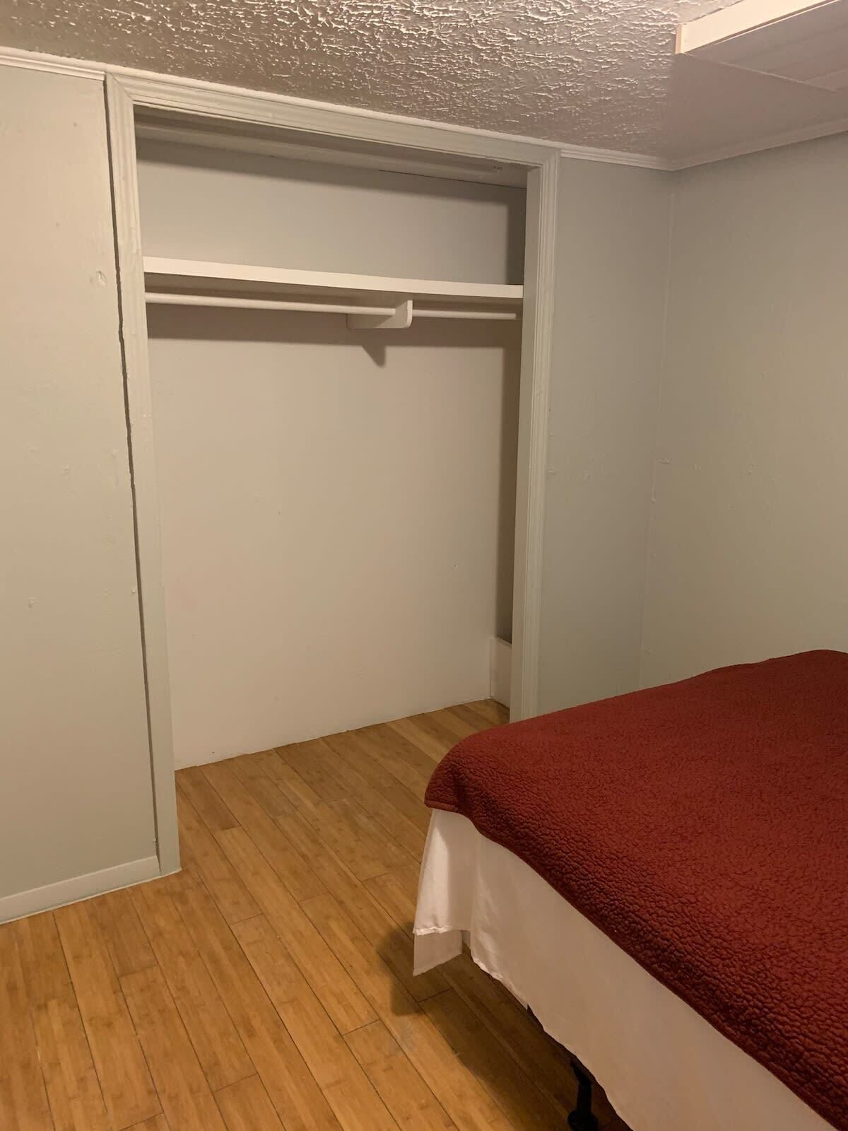Ein Schlafzimmer mit Holzboden, einem Bett mit roter Decke und einem weißen Kleiderschrank mit Kleiderstange.