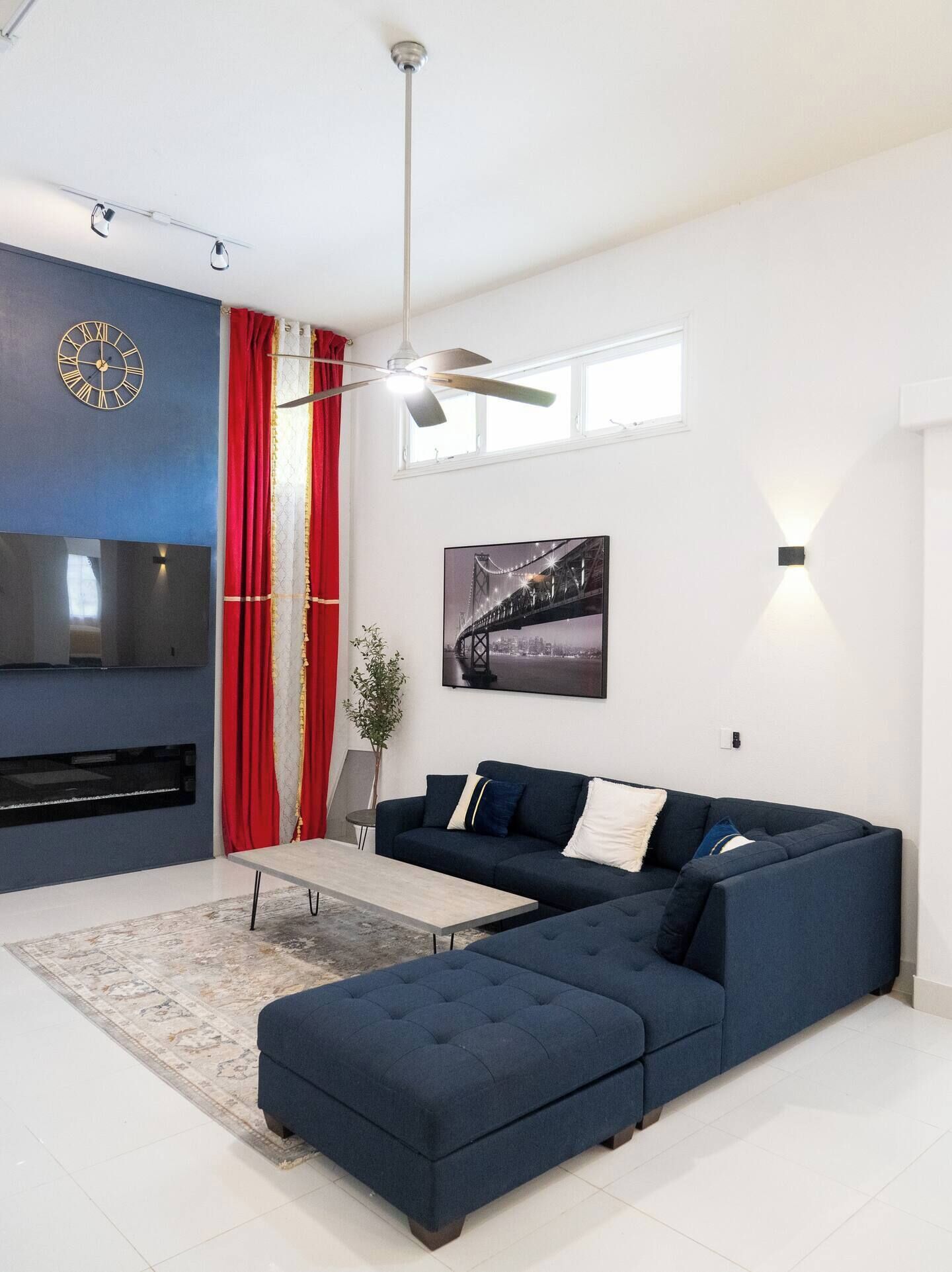 A modern living room with a sectional sofa, a flat-screen TV, a ceiling fan, and a large window.