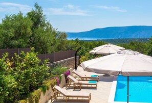 A poolside area with lounge chairs and umbrellas, surrounded by greenery and overlooking a body of water with mountains in the distance.