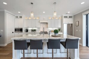 Uma cozinha moderna com armários brancos, uma ilha de mármore, quatro banquetas altas, eletrodomésticos de aço inoxidável e iluminação pendente.