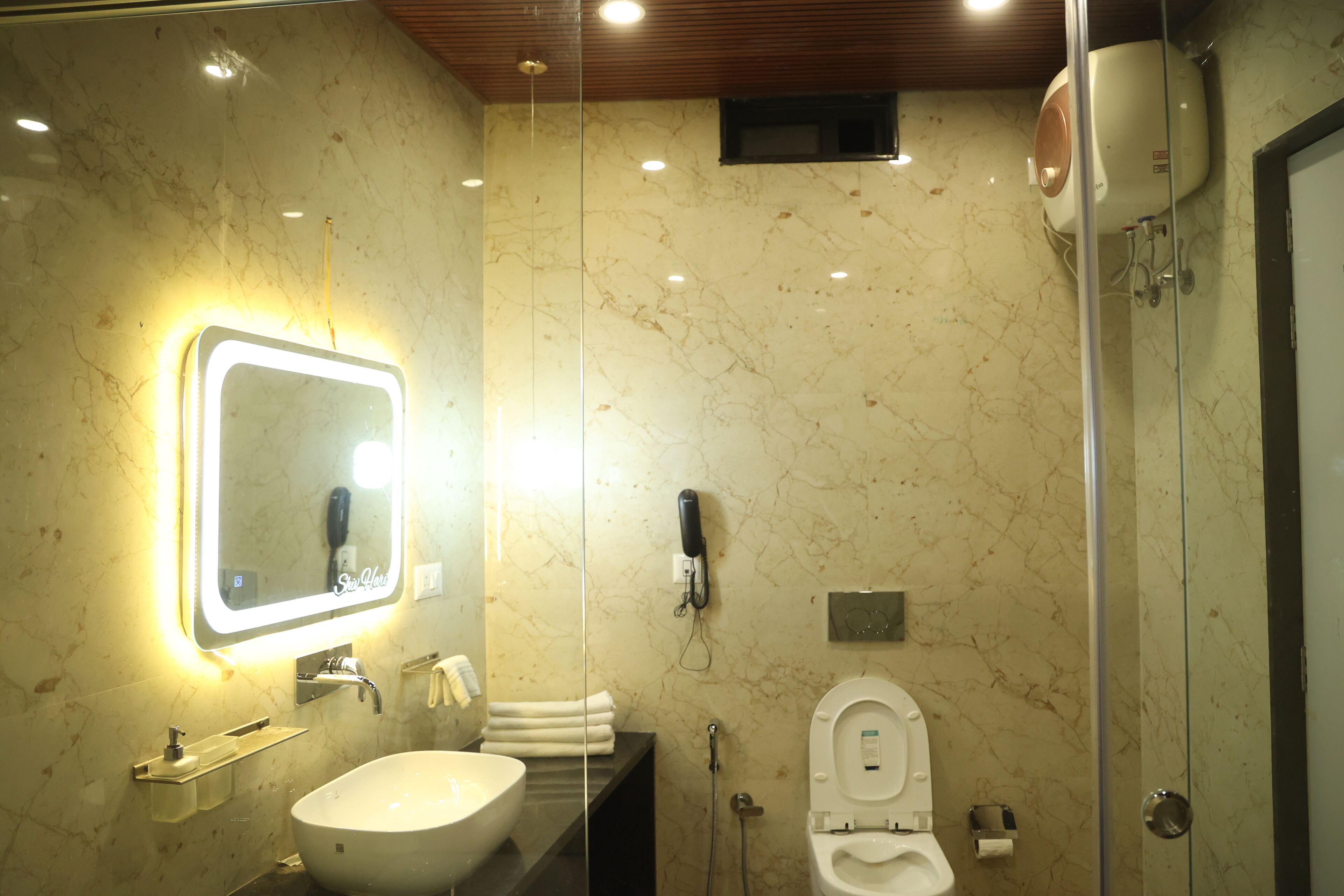A modern bathroom with a marble wall, a large sink, a mirror with lights, a toilet, and a shower.