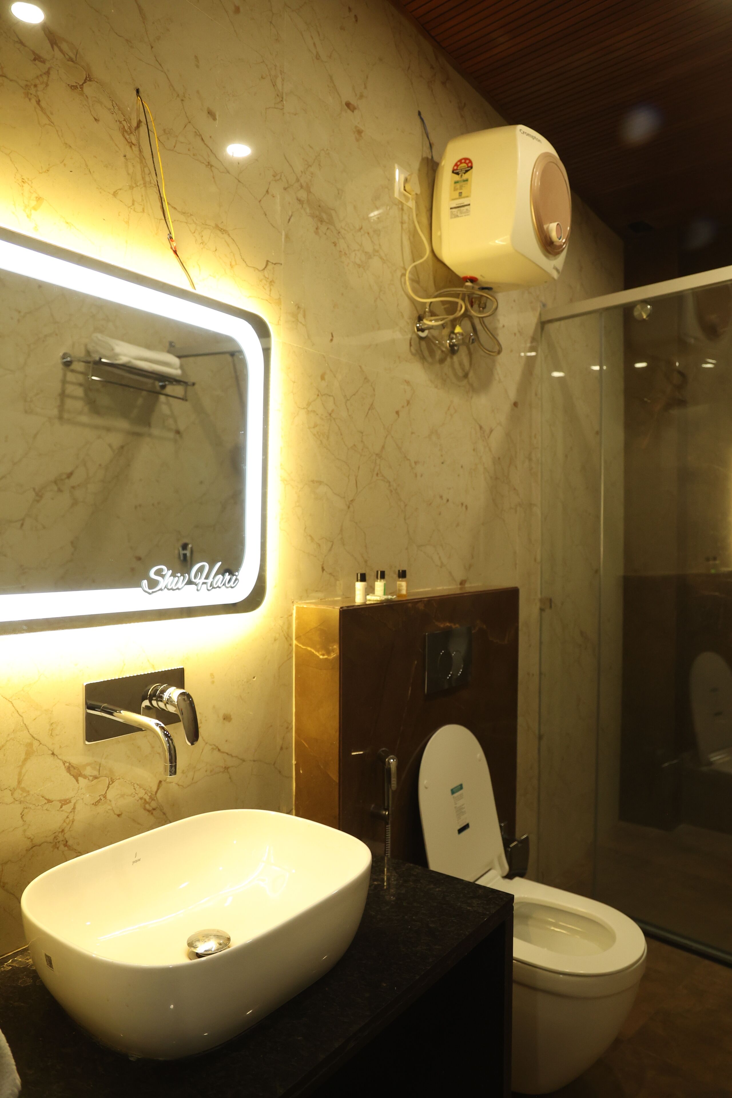 Un baño con pared de mármol, lavamanos blanco, espejo con luz, inodoro y zona de ducha.
