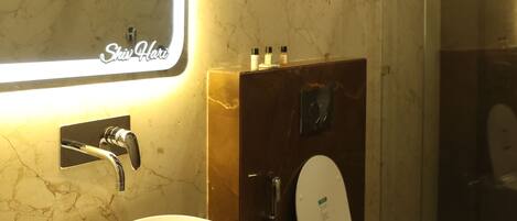 Un baño con pared de mármol, lavamanos blanco, espejo con luz, inodoro y zona de ducha.