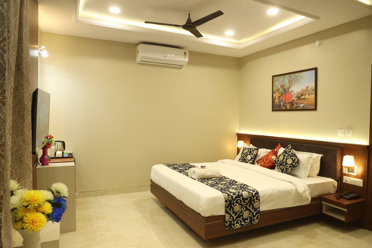 A hotel room with a bed, bedside table, nightstand, lamp, air conditioner, ceiling fan, and a painting on the wall.