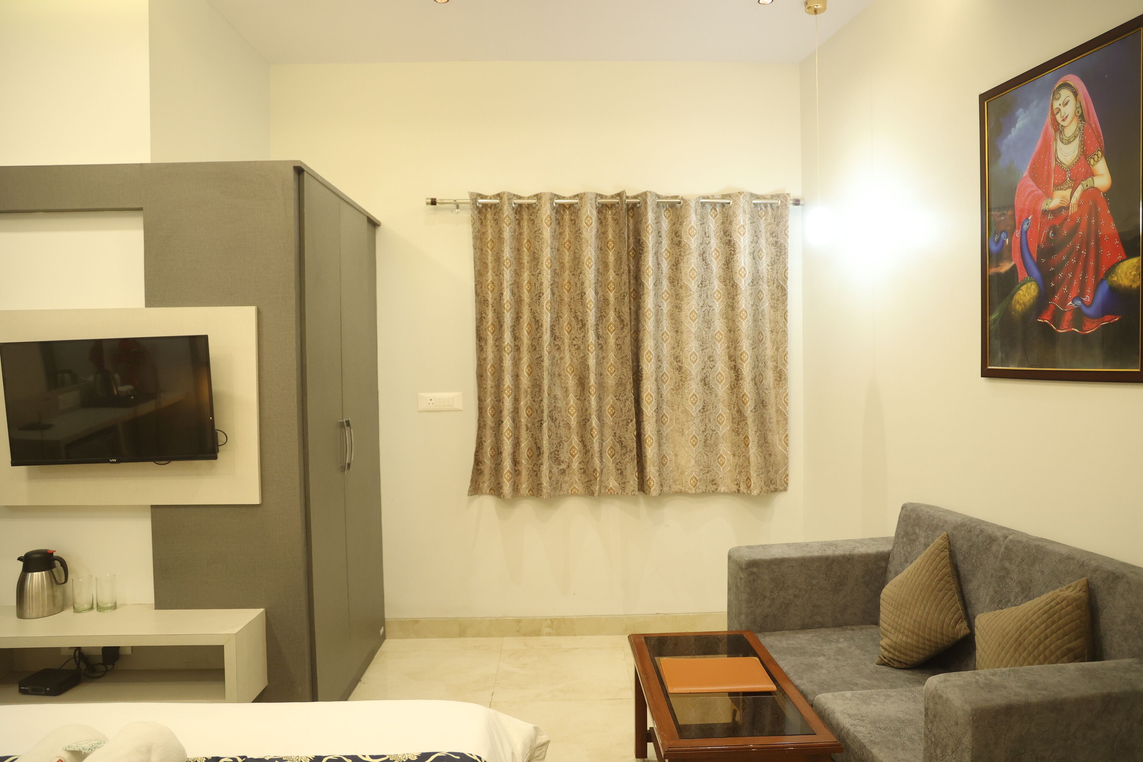 A living room with a grey sofa, a small wooden coffee table, a flat-screen TV mounted on a white cabinet, a patterned curtain, and a framed artwork on the wall.