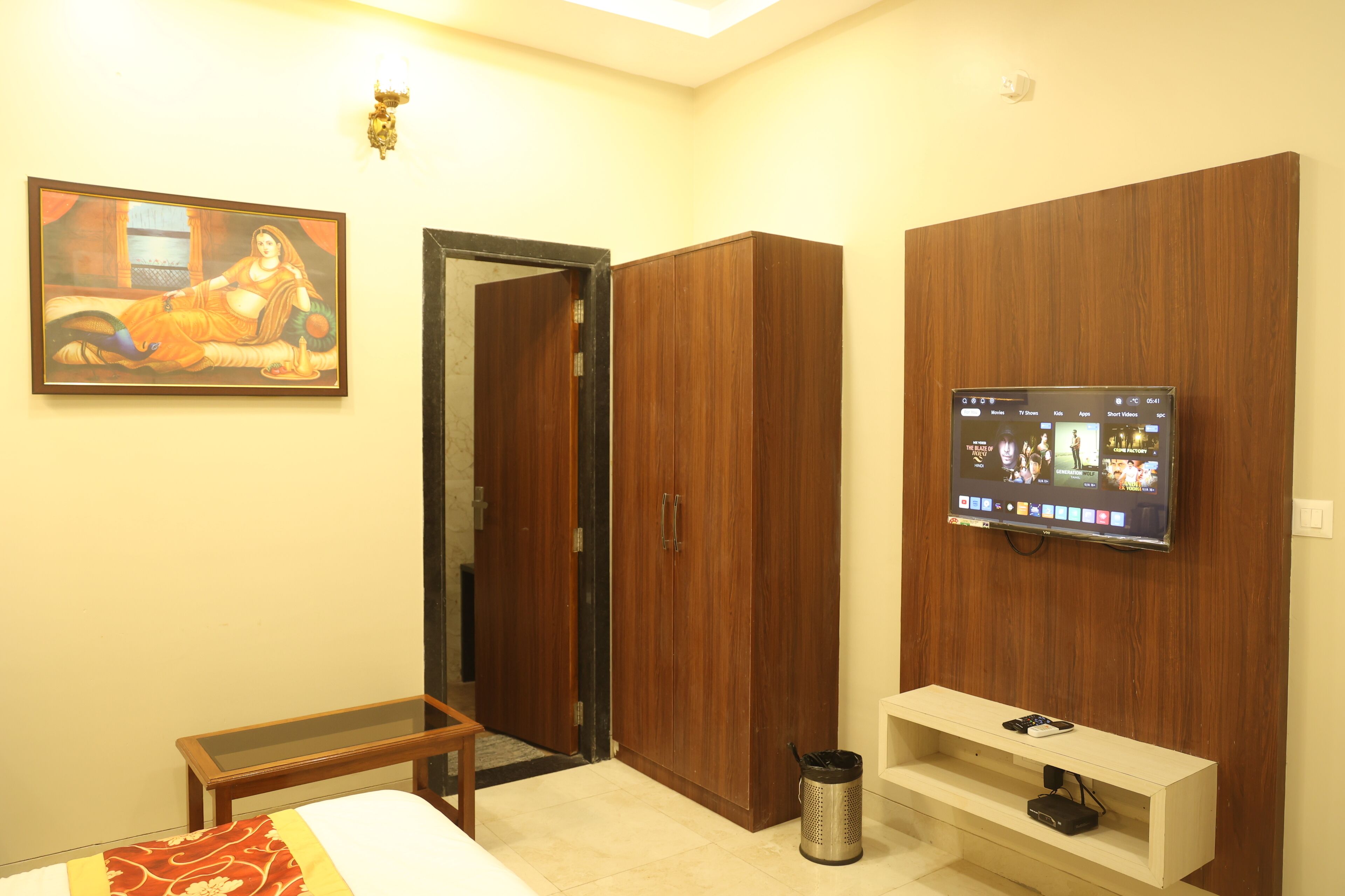 A hotel room with a bed, a TV mounted on the wall, a wooden wardrobe, and a painting on the wall.