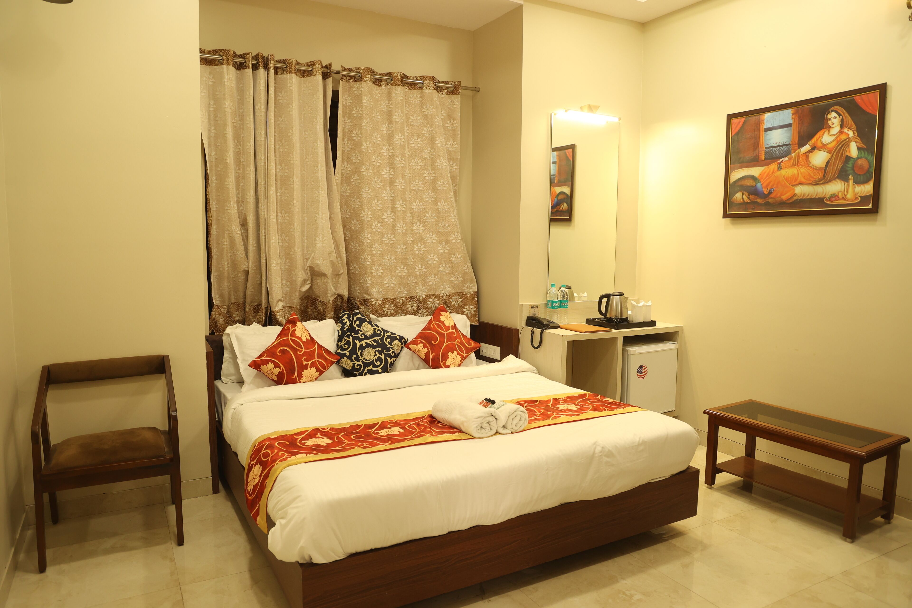 A neatly made bed with a red and gold bedspread, a wooden nightstand, a small table, a chair, and a painting on the wall.