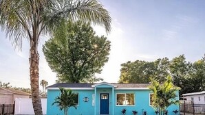 Ein eingeschossiges Haus mit blauer Fassade, einer Haustür und einem kleinen Vorgarten mit einer Palme und Blumentöpfen.