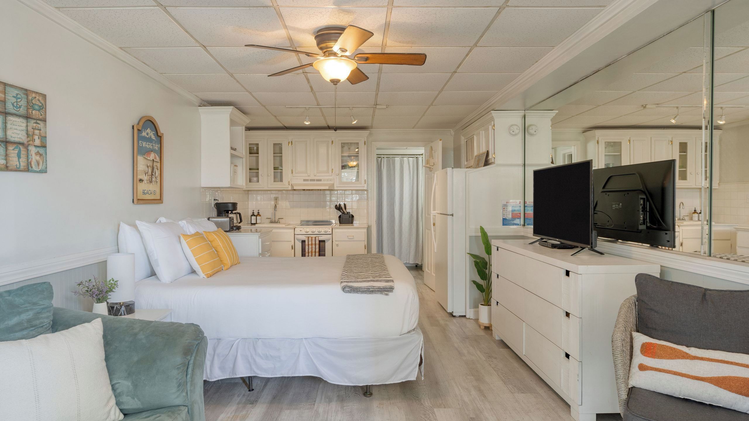 A bedroom with a bed, a ceiling fan, a TV, and a kitchenette in the background.