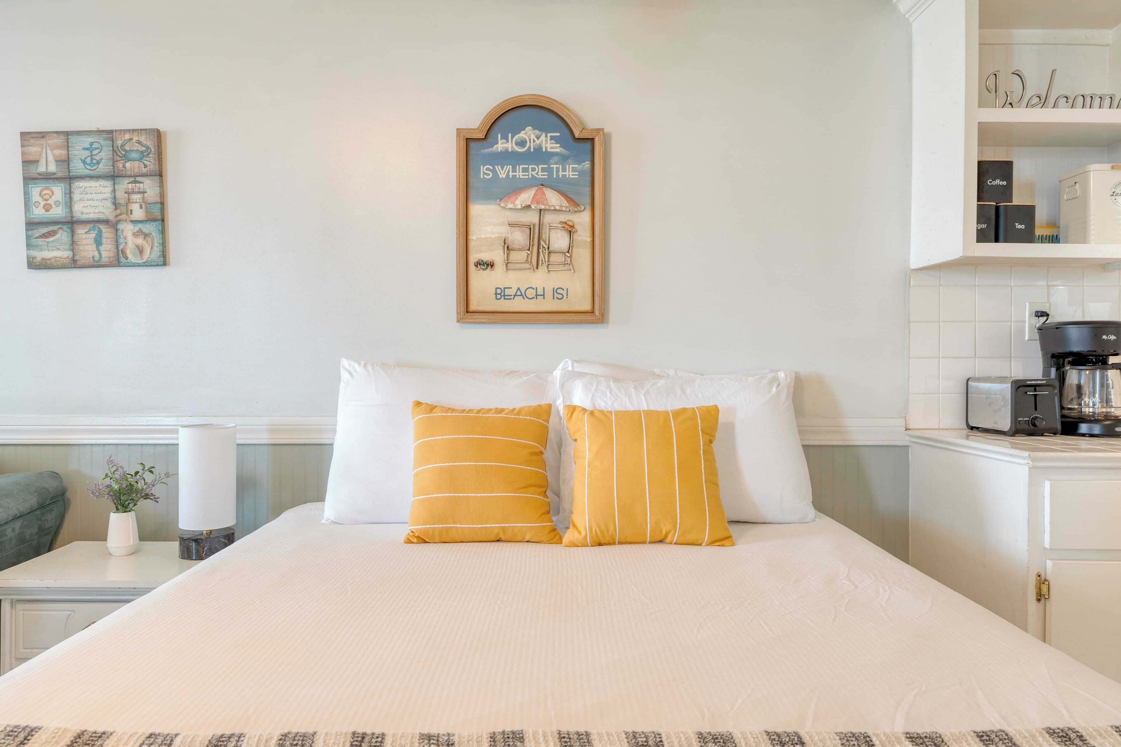 Una cama bien hecha con almohadas a rayas blancas y amarillas, un cartel enmarcado encima de la cama y una cafetera sobre una encimera a la derecha.
