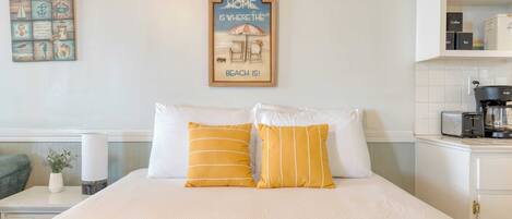 Una cama bien hecha con almohadas a rayas blancas y amarillas, un cartel enmarcado encima de la cama y una cafetera sobre una encimera a la derecha.