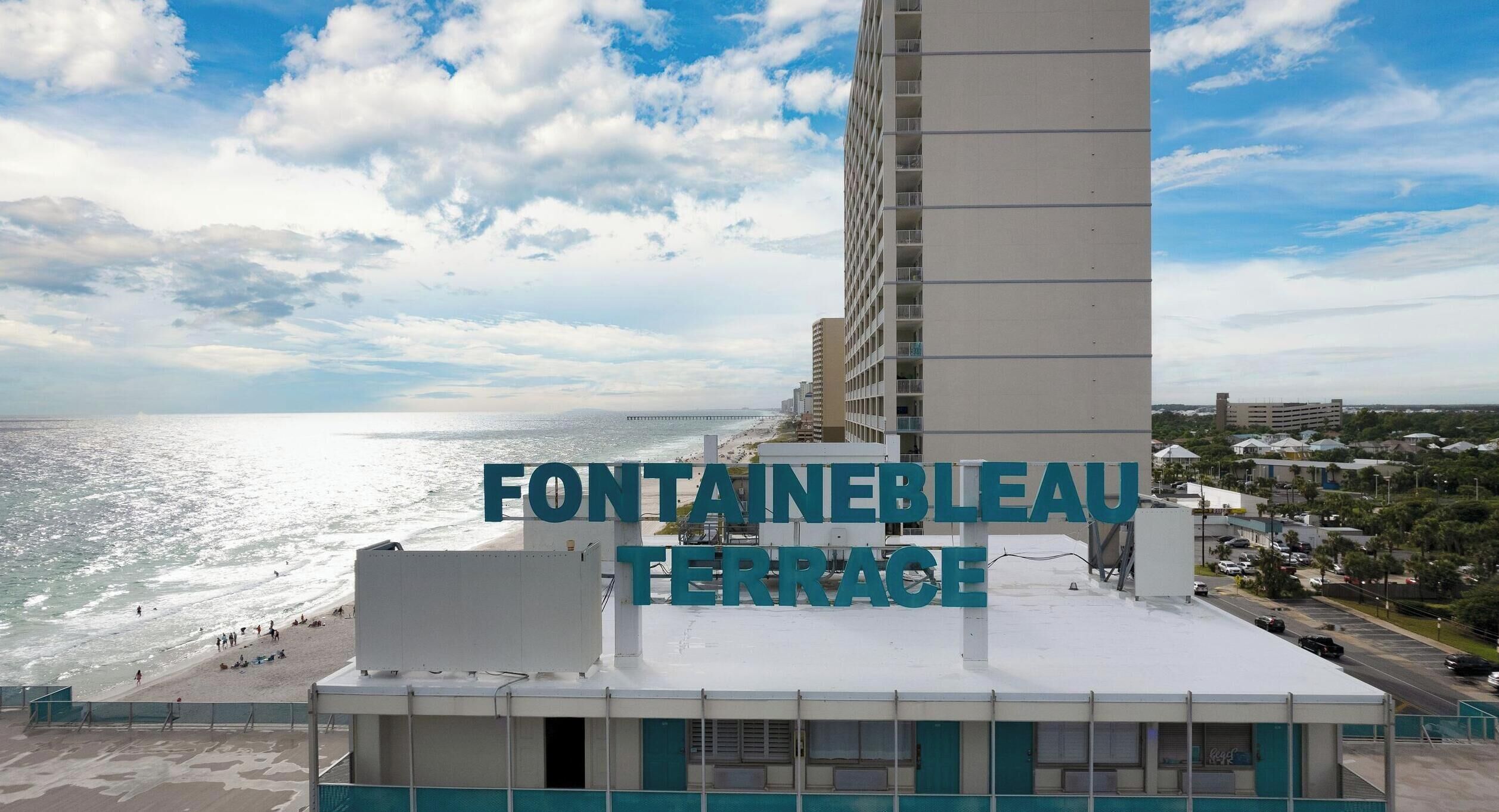 A high-rise hotel with a prominent name sign, a beach view, and a parking area.