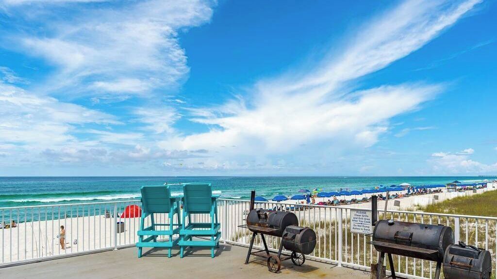 A balcony with two chairs, a grill, and a view of the beach and ocean.