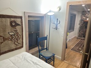 A bedroom with a bed, a chair, a wall-mounted lamp, and a door leading to a stone fireplace.