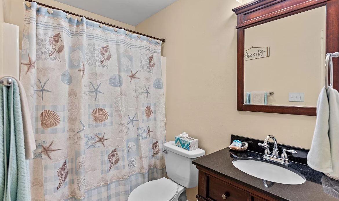 A bathroom with a shower curtain featuring sea shells and starfish, a toilet, a sink with a mirror, and a towel rack.
