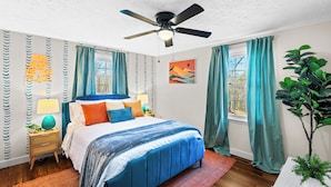 Una camera da letto con un letto, una ventola a soffitto, una finestra con tende, un comodino con una lampada e una parete con un cartello decorativo.