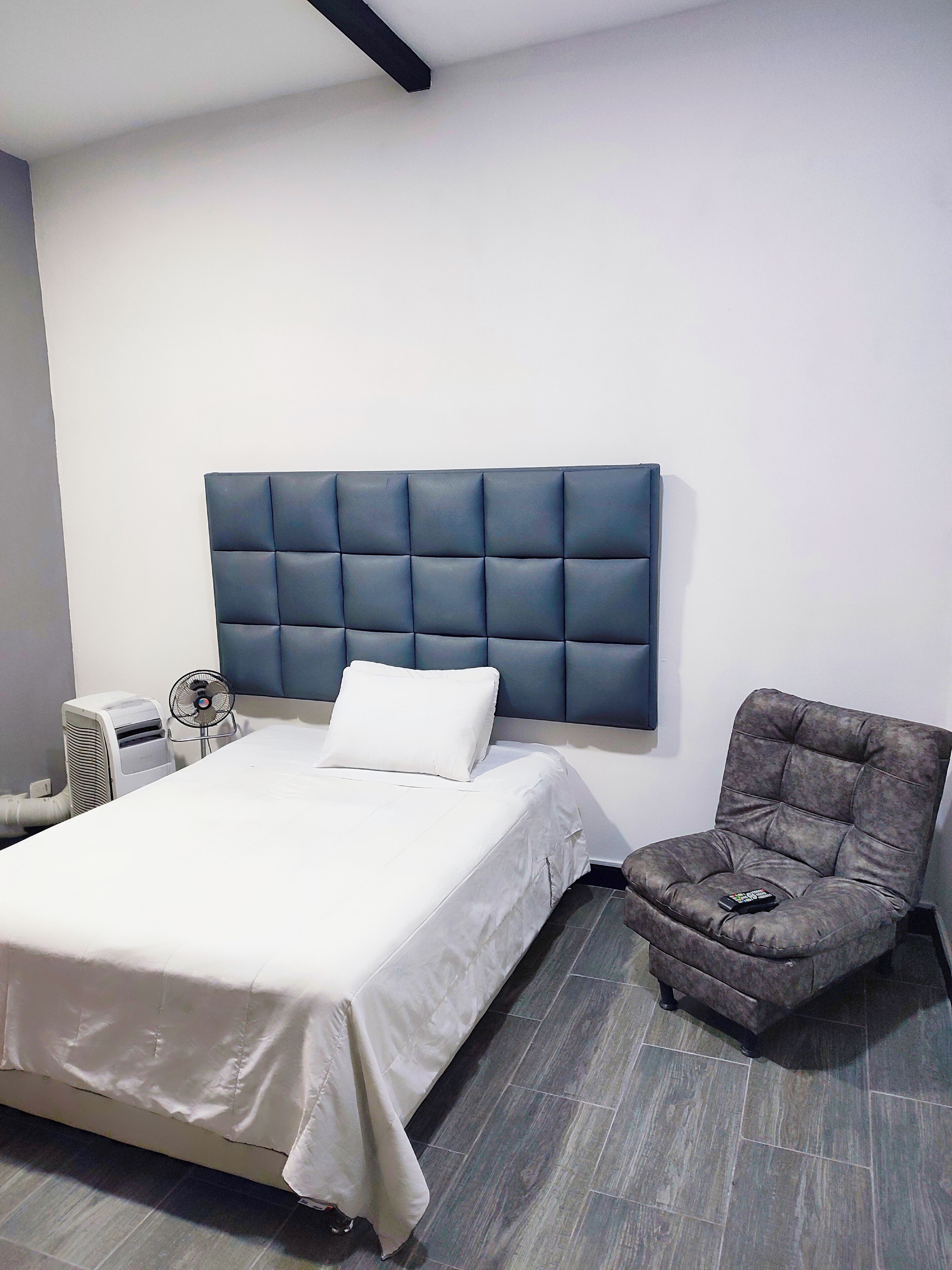 Une chambre moderne avec un grand lit, un fauteuil à revêtement gris, une table de chevet avec un ventilateur et un climatiseur fixé au mur.