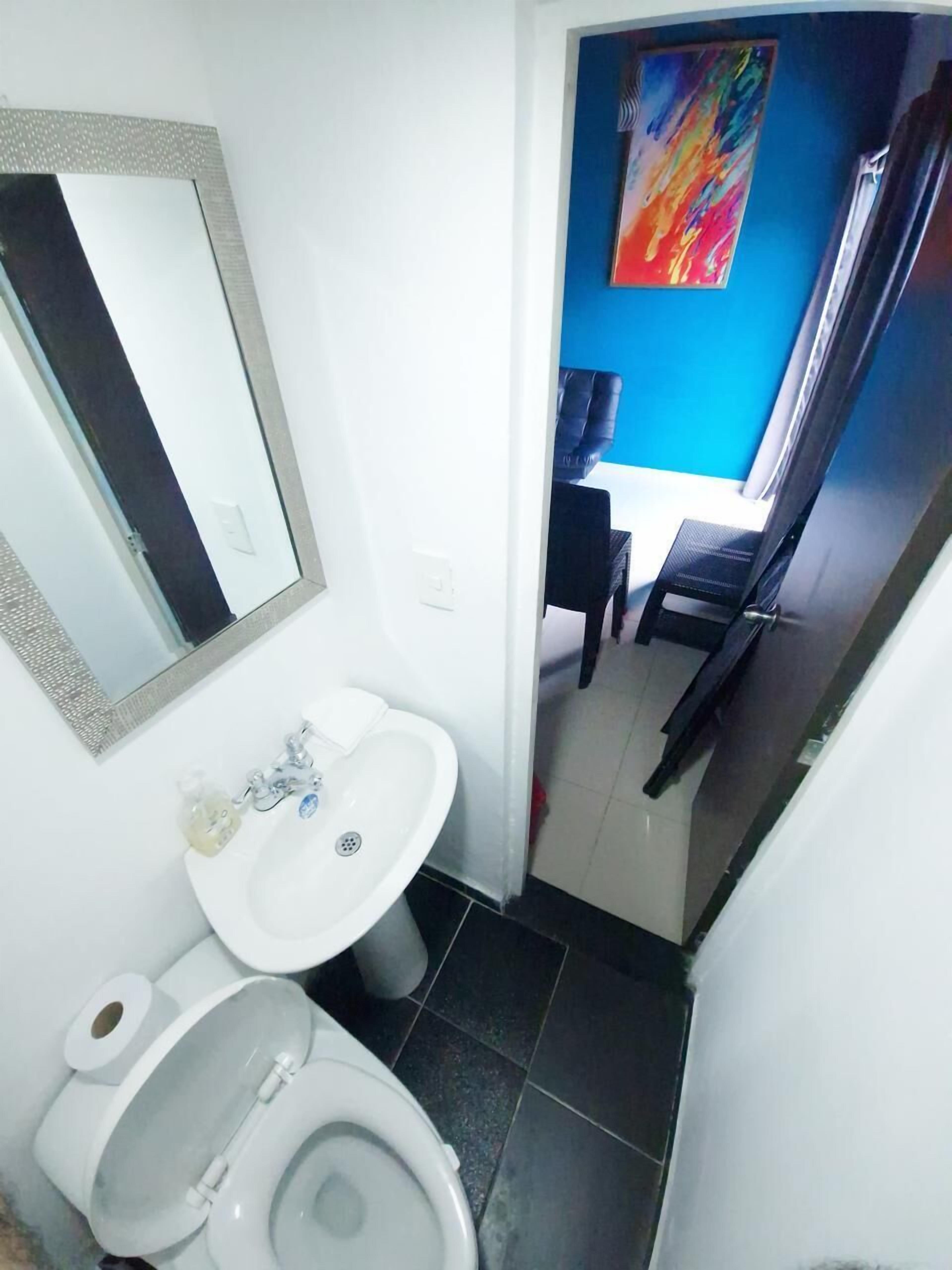 A small bathroom with a white sink, a toilet, and a mirror. There is a blue wall and a painting visible in the background.