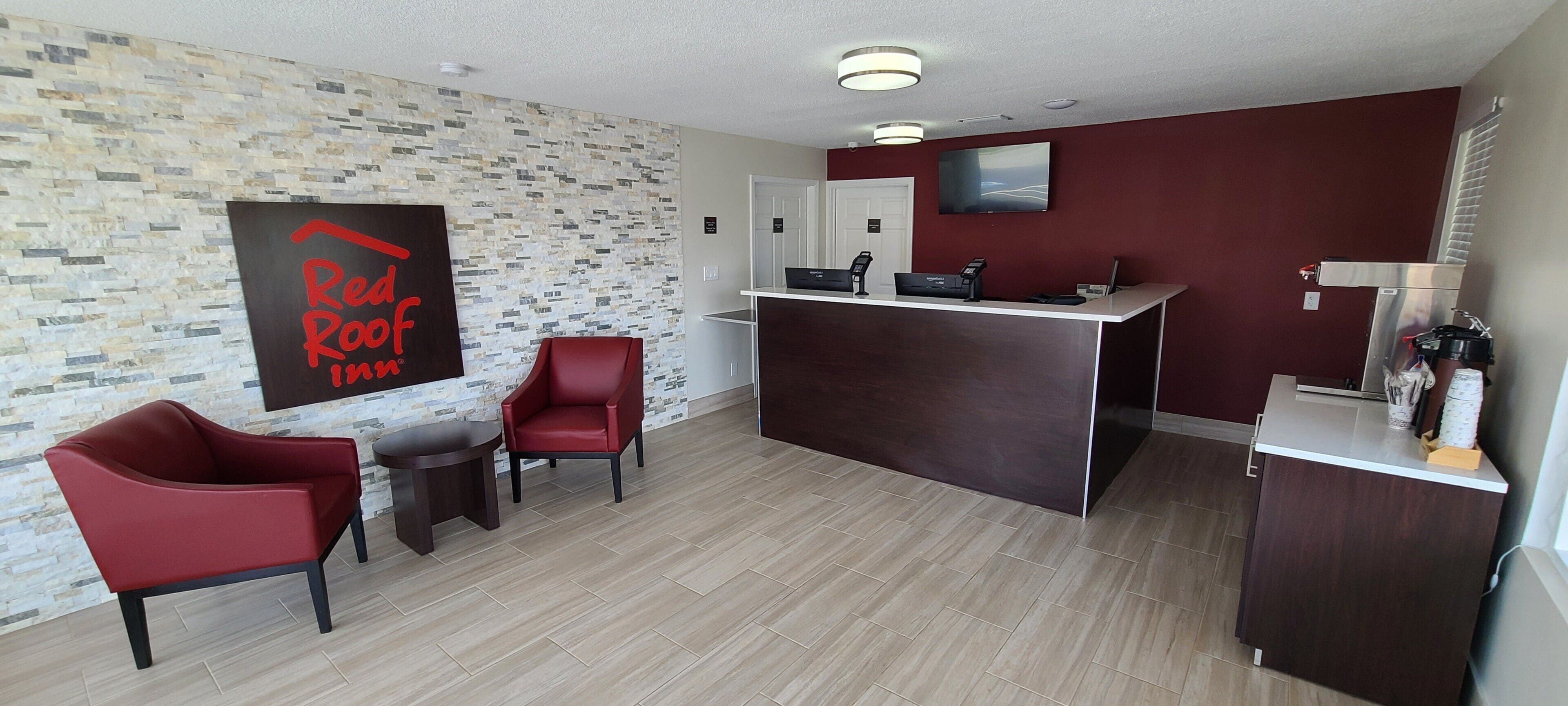 A Red Roof Inn reception area with a modern design, including a reception desk, two red chairs, a small table, and a wall-mounted TV.