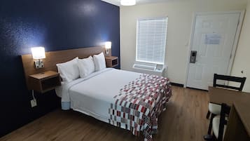 Quarto de hotel com uma cama, mesas de cabeceira com abajures, uma janela com persianas, uma porta e uma pequena mesa com uma cadeira.
