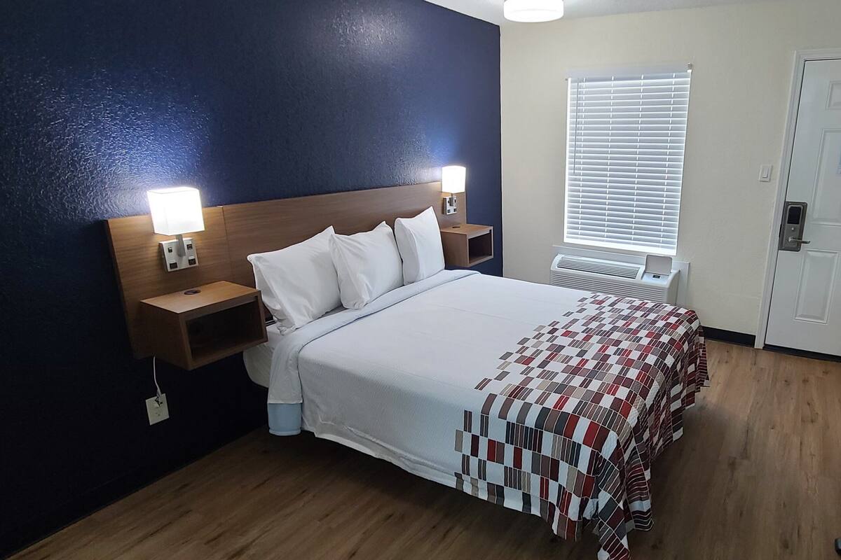 A hotel room with a bed, bedside tables with lamps, a window with blinds, a door, and a small table with a chair.
