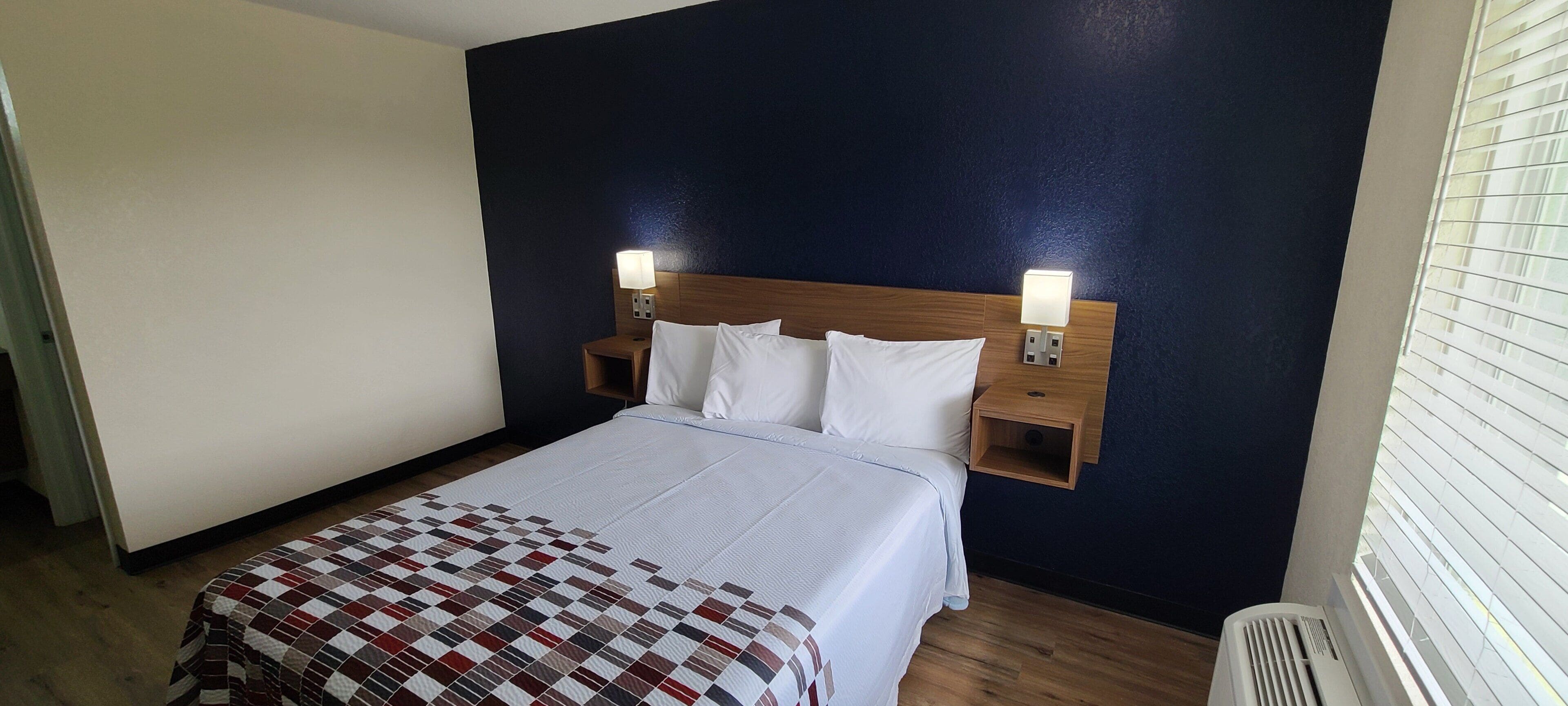 Ett hotellrum med en säng, två sänglampor, en träpanel vid sänggaveln och ett fönster med persienner.