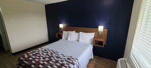 A hotel room with a bed, two bedside lamps, a wooden headboard, and a window with blinds. - Red Roof Inn Byron (Byron)