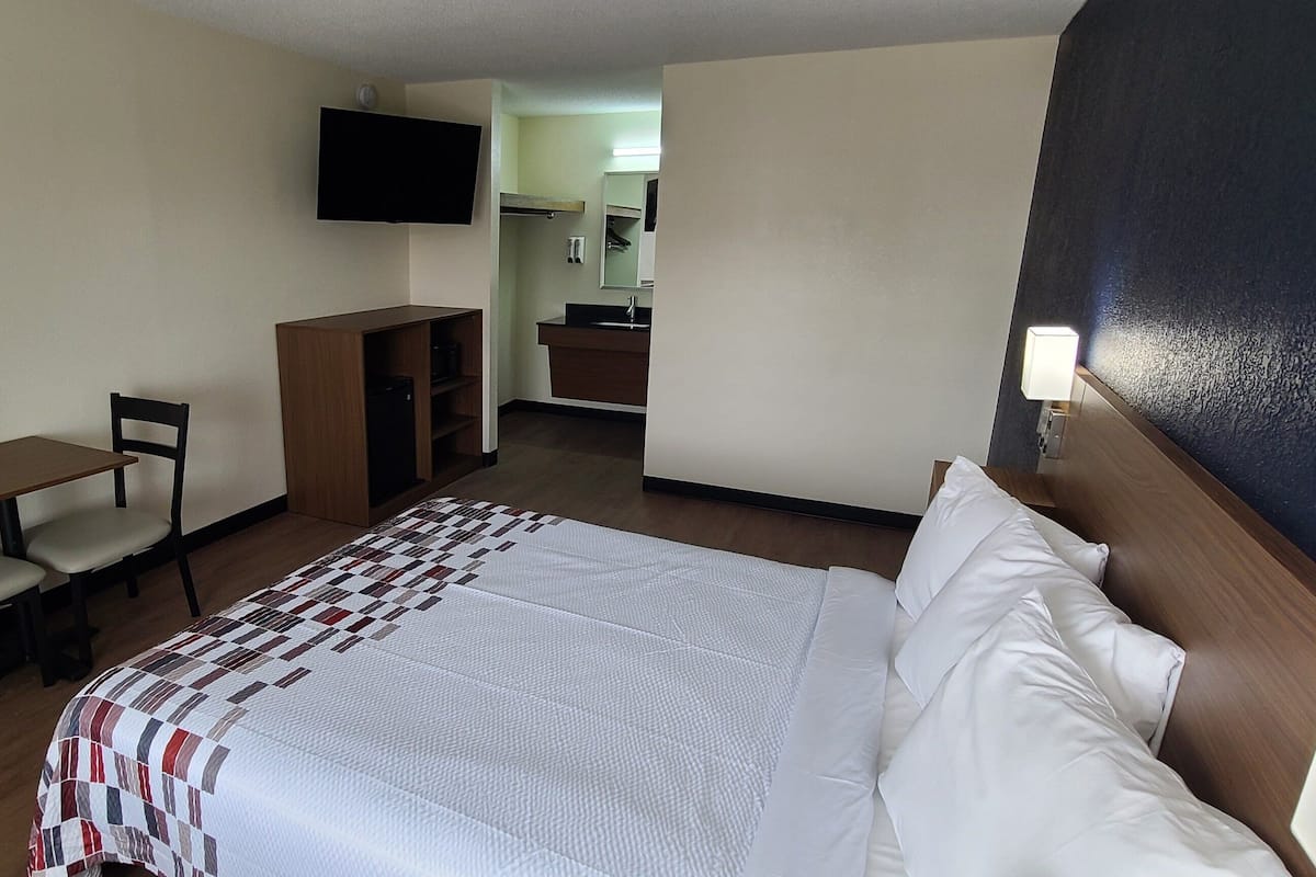 A hotel room with a bed, a desk, a chair, a TV, and a bathroom visible through an open door.