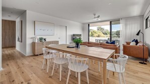 Un salon moderne avec une table à manger en bois, des chaises blanches, un canapé marron et une grande fenêtre donnant sur l’extérieur.