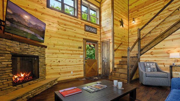 A log cabin interior with a stone fireplace, a mounted TV, a staircase, and a cozy seating area. - Mountain Magic: The Perfect Romantic Cabin Getaway (Gatlinburg)