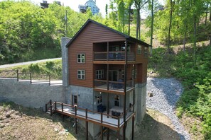 A two-story wooden cabin with a balcony on the upper floor, surrounded by trees and a gravel path.