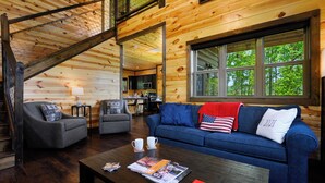 A wooden cabin interior with a living area featuring a blue sofa, two armchairs, a coffee table with magazines, and a large window overlooking greenery. - Mountain Magic: The Perfect Romantic Cabin Getaway (Gatlinburg)