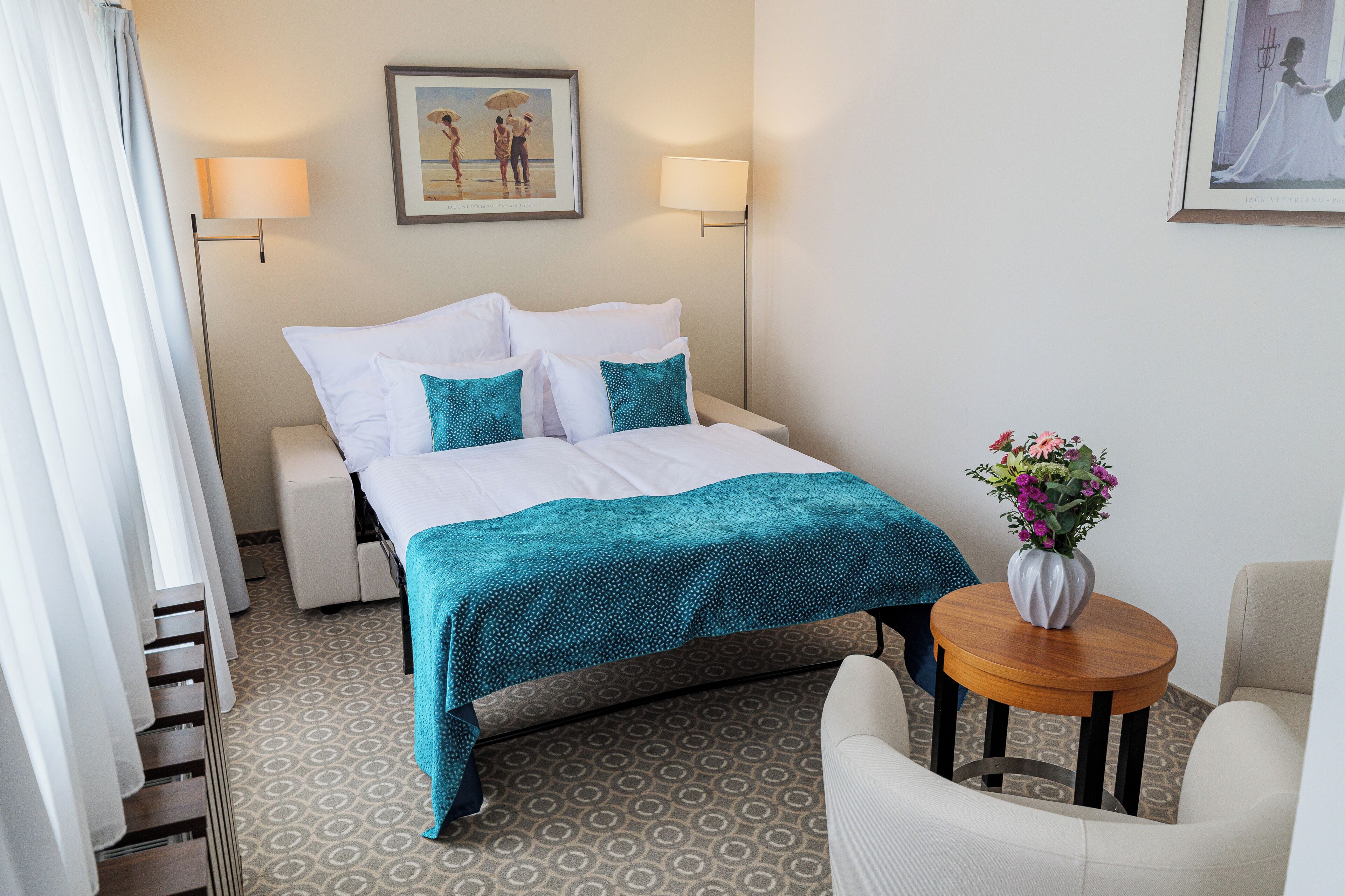 Quarto de hotel com cama, abajures, uma mesinha com um vaso de flores e uma cadeira.