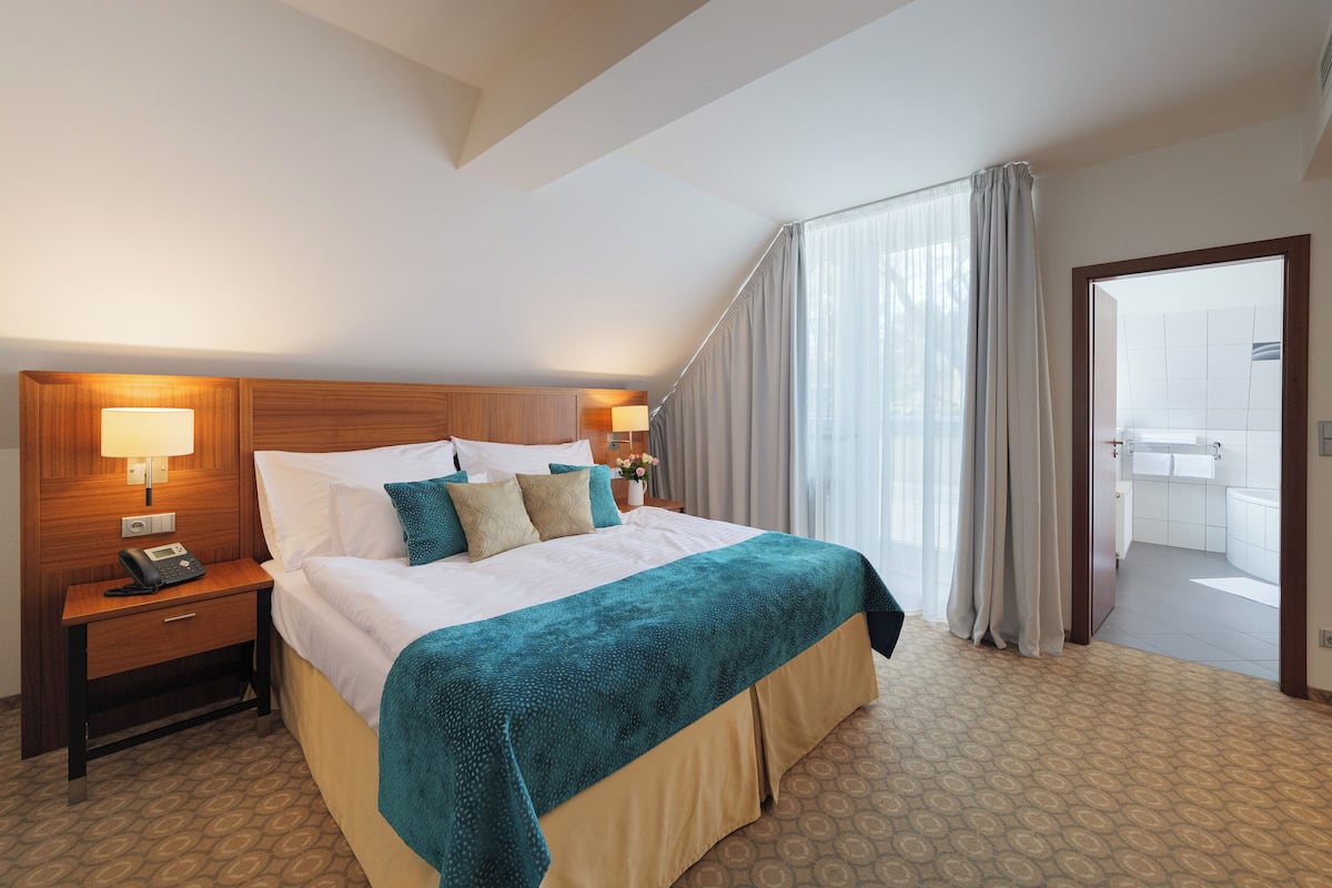 A hotel room with a large bed, a bedside table with a lamp, and a view of the balcony through the window.