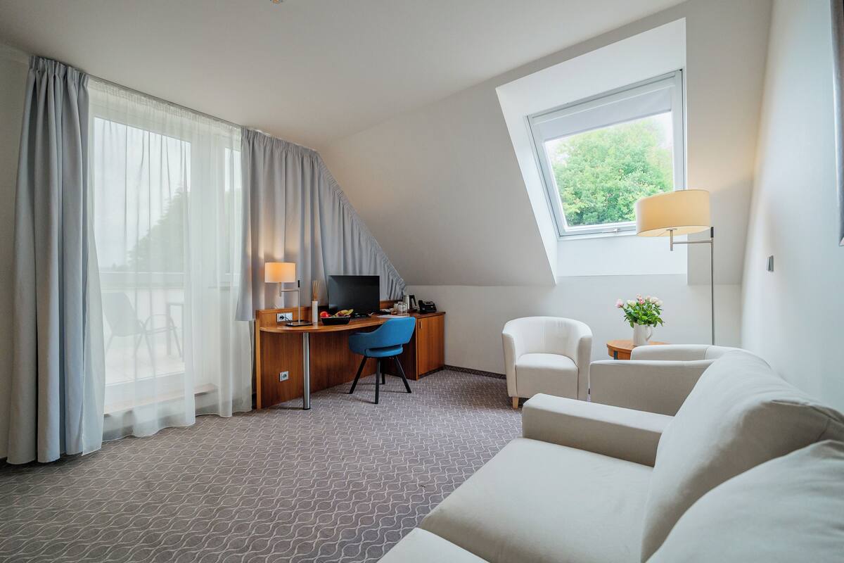 A modern hotel room with a desk, a blue chair, a white sofa, and a large window with a view of greenery.