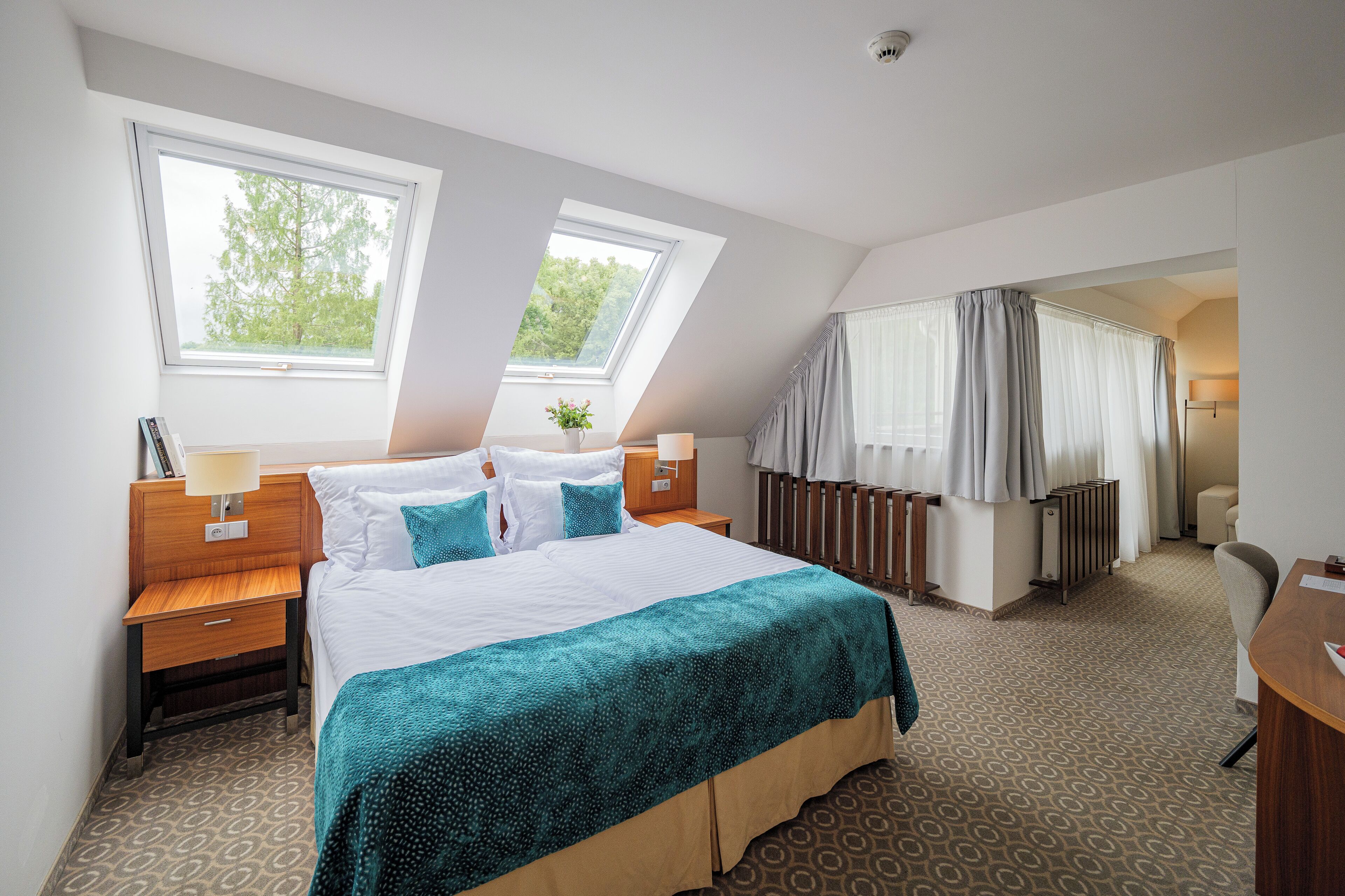 Habitación de hotel con una cama grande, dos ventanas, una mesita de noche, una lámpara, un radiador y una silla.