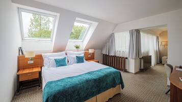 Habitación de hotel con una cama grande, dos ventanas, una mesita de noche, una lámpara, un radiador y una silla.