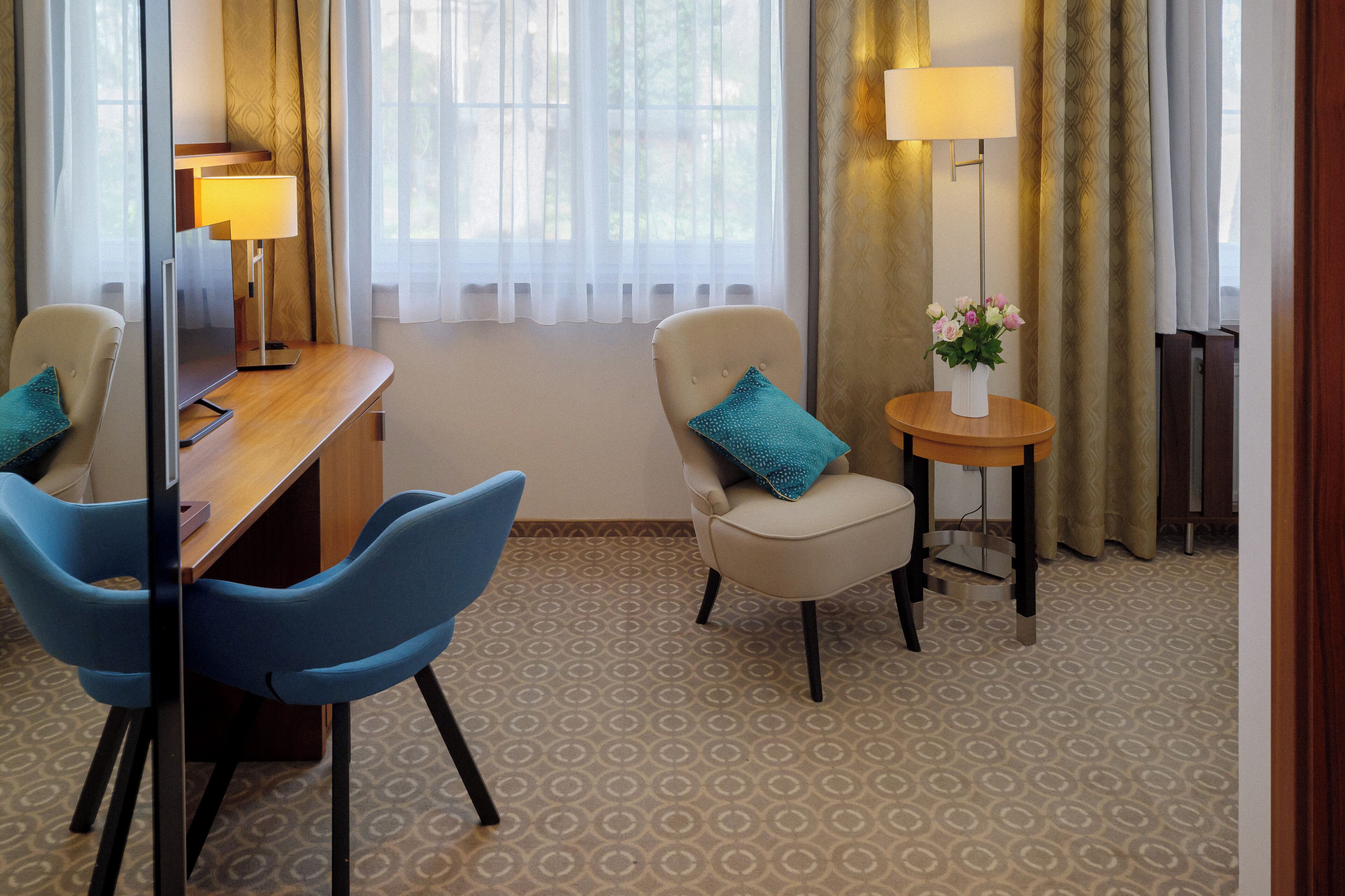 Une chambre d’hôtel avec un bureau en bois, deux chaises, une petite table avec une lampe et un vase rempli de fleurs.