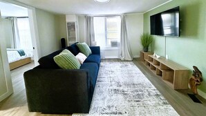A living room with a sofa, a flat-screen TV, a potted plant, and a window with curtains. - Charming & Bright Gem: Awesome Location/AC/Laundry (Renton)